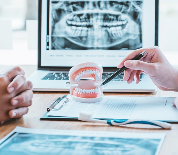 a pen pointing to a model of a mouth and a laptop displaying X-rays in the background